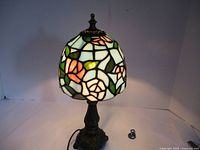 Tiffany style lamp illuminated, showing colorful stained glass floral design on shade and bronze-colored ornate base.