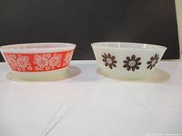 Two vintage Anchor Hocking glass bowls side by side on a flat surface against a dark background. One bowl has brown daisy flower prints, the other has an orange-red floral pattern.