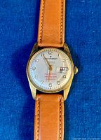 Front view of the Star Master wrist watch showing the silver dial, Arabic numerals, gold-tone hands, date window, and light brown leather strap.