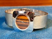 Front angled view of the vintage ladies watch showing the tiger's eye dial, polished chrome square case, and wide chrome bracelet against a blue background.