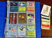 Blue binder open to show 9 Pokemon cards in plastic sleeves. Cards include holographic Darkrai, Abomasnow, Vulpix, and others plus a stack of assorted cards to the side.
