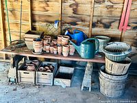 Complete view of the gardening items arranged on and under a wooden workbench against a wood paneled wall including terra cotta pots, wooden planters, watering cans in blue and green, and metal garden supports.