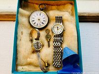 Four watches laid in padded box: silver pocket watch, Birka watch, Bella watch, brass key
