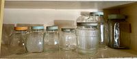 Shelf display of seven antique glass jars with metal lids of varying sizes and designs.