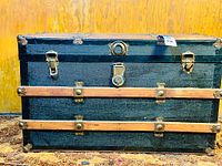 Front view of the antique steamer trunk showing black surface, wooden slats, original brass locking mechanism, and side latches.
