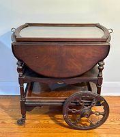 Full view of the antique wooden bar cart showing drop-leaf top in folded position, lower shelf, and large decorative metal wheels.