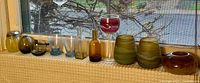 Photo of 12 various art glass cups and small vessels on a windowsill, showing colors and shapes.