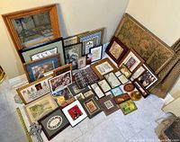 Photo shows a collection of framed art arranged on floor including prints, varying sizes, a large wooden framed mirror, and decorative framed artworks in a corner of a room.