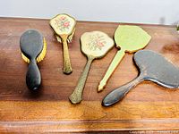 Five vanity items on wood surface showing two floral hand mirrors with ornate handles, one green-handled mirror, and two black brushes.