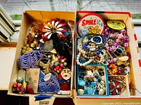 Wide view of assorted estate jewelry collection in two open boxes on window sill showing colorful beads, cluster earrings, wooden and metal brooches, and pinback buttons.