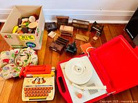 Photo showing various vintage toys including the Tutor Typer toy typewriter, box of plastic pretend food, fabric hot dog toy, various wooden dollhouse furniture, black toy sewing machine, and red and white portable record player opened.