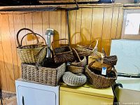 Big collection of woven baskets of various sizes and shapes placed on a white dryer and a yellow washing machine against a wood-paneled wall.