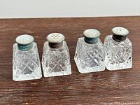 Four salt and pepper shakers with sterling silver tops and mother of pearl lids displayed side by side on a flat surface, showing cut crystal glass bodies and the different shaped holes for salt and pepper dispensing.