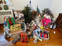 Wide view showing assorted Christmas decorations including a small artificial tree, ornaments in boxes and bags, nativity figures, Santa village houses, and various other décor items grouped on the floor.