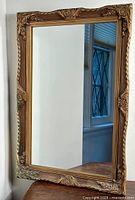 Front view of entire rectangular mirror leaning against wall on table, showing the baroque style ornate frame with floral and scroll decorative details in brown-gold finish.