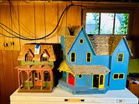 Wide shot showing both handmade wooden dollhouses, one blue with yellow window frames and one Victorian style with natural shingles and red porch