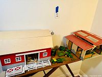 Photo showing red and white wooden barn, beige stable with red roof, and multiple green farming toy tractors and equipment on table