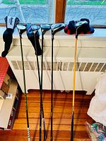 Full view of six golf clubs leaning against radiator, some with black and multicolor socks/headcovers.