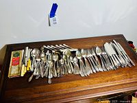 Overview of entire flatware and cutting utensil collection laid out on a wooden table showing various spoons, forks, knives and boxed utensils.