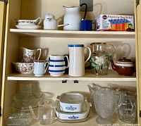 Three shelves inside a wooden cabinet with various kitchen items including multiple pitchers, Corelle dishes, and glassware.