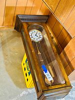 Full view of large wood regulator wall clock with glass front showing clock face and pendulum.