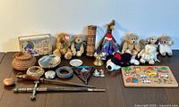 Wide photo capturing the entire assortment of assorted vintage items laid on a wooden table including teddy bears, baskets, miniature sword, chocolate box, bell, puzzle board and others