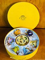 Top view of round yellow box with six butterfly-themed tea cups and saucers arranged inside on white satin lining.