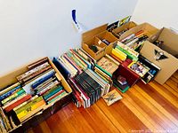 Wide view of multiple boxes filled with a large variety of books arranged by size and genre on hardwood floor