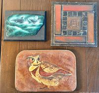 Three pieces of mid-century wall art displayed on a wooden floor showing the owl enamel on copper, geometric tile, and green bird enamel piece.