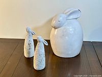 Three minimalist bunny rabbit sculptures displayed on a wooden surface: one large smooth glazed bunny and two smaller textured bunnies.