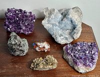 Overview photo showing all seven semi-precious gemstone clusters arranged on a wooden surface. Includes two amethyst clusters, one celestite, malachite+azurite, emerald/quartz, and two mica clusters.