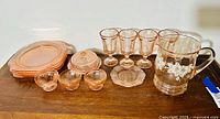 Pink depression glass set comprised of 4 square plates, 3 small bowls, 3 small plates, a covered butter dish, 5 goblets, and a clear pitcher with floral decoration arranged on wooden surface.