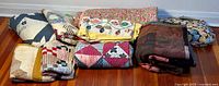 Group photo showing 7 folded quilts and patchwork covers in assorted patterns and colors laid in a row on the floor.