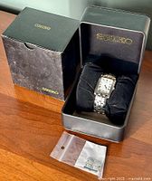 Seiko men's wristwatch resting inside a black Seiko metal box with original outer black cardboard sleeve. Extra metal links in a clear plastic bag are placed in front of the box on a wooden surface.