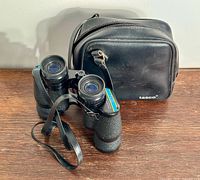 TASCO binoculars and original black case side by side on a wooden surface, showing textured body, shoulder strap, and case details