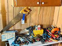 Wide view of counter with various hand tools including saws, pliers, hammers, knives, tape measure, and a staple gun.