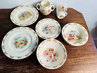 Full view of 6-piece set of vintage Royal Doulton Bunnykins dishes on wooden table, showing plates, bowls, and handled mugs with bunny motifs and border detail
