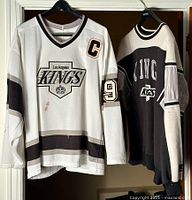 Front and side view of the two garments: Wayne Gretzky white CCM LA Kings hockey jersey with captain patch and number 99 on sleeve, alongside black Starter LA Kings pullover sweater, both hanging on hangers