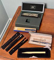 Full view of vintage pens on wooden table showing cases including Cross set, yellow/blue ballpoint, Waterman fountain pen, and assorted pens in velvet sleeves.