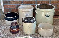 Photo of six vintage stoneware crocks of varying sizes arranged on a tiled floor against a brick wall, showing different colors, markings, and wear.