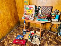 Wide shot showing the table with all vintage board games, puzzles and toys piled on top including a Pictionary box, electronic toys, and chess/checkers set.