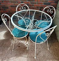 Angled view showing round glass-top table with white wrought iron scrollwork frame and four matching chairs with teal cushions.