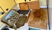 Wide shot showing multiple wooden cutting boards stacked, Florentine Italy metal tray with intricate designs, wicker tray with cottage scene, wooden rolling pin, wire toast rack, and a wire cooling rack.