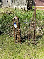 Two wrought iron garden obelisks shown outdoors on grass, one tall and pointed, one smaller rectangular cuboid