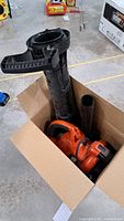 Leaf blower unit with orange body and black handle placed inside a cardboard box, showing the blower tube and vacuum components.