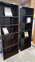 Two tall black wooden book shelves with adjustable shelves, holding four decorative picture frames.