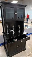 Front view of black buffet cabinet showing glass upper doors, open shelf with three silver candlesticks, lockable drawer pulled open, and closed lower doors.