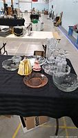 Overview of glassware items on black tablecloth including trays, vase, bowls, flutes, and two decorative dolls