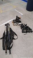 Two aluminum baseball bats in black and silver colors placed beside a black soft carrying bag, two pairs of roller blades with one pair black and grey men's size 11 and the other smaller size in black with purple accents.