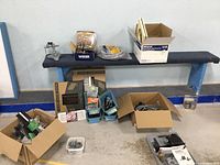 Overall view of multiple cardboard boxes on and under a bench containing mixed hardware items including door handles, registers, and plumbing parts.